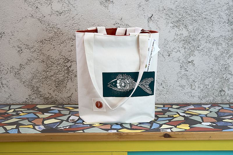 Natural canvas tote with a deep teal printed panel showing a hand-carved fish and eye, lined in rust orange.