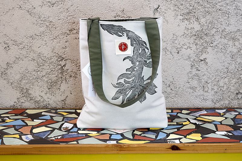 Pale canvas tote with olive webbing straps, printed with a fine grey plume motif and the workshop's oxblood seal.